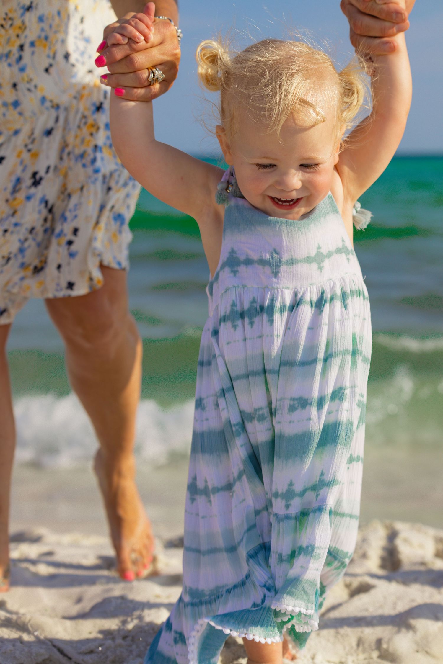 Toddler in tie-dye outfit plays on sunny beach with waves in background.