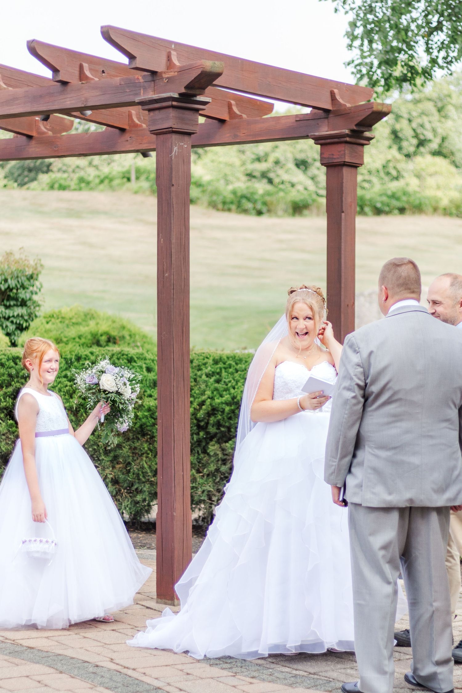 Wedding ceremony under wooden pergola with bride in white dress and flower girl.