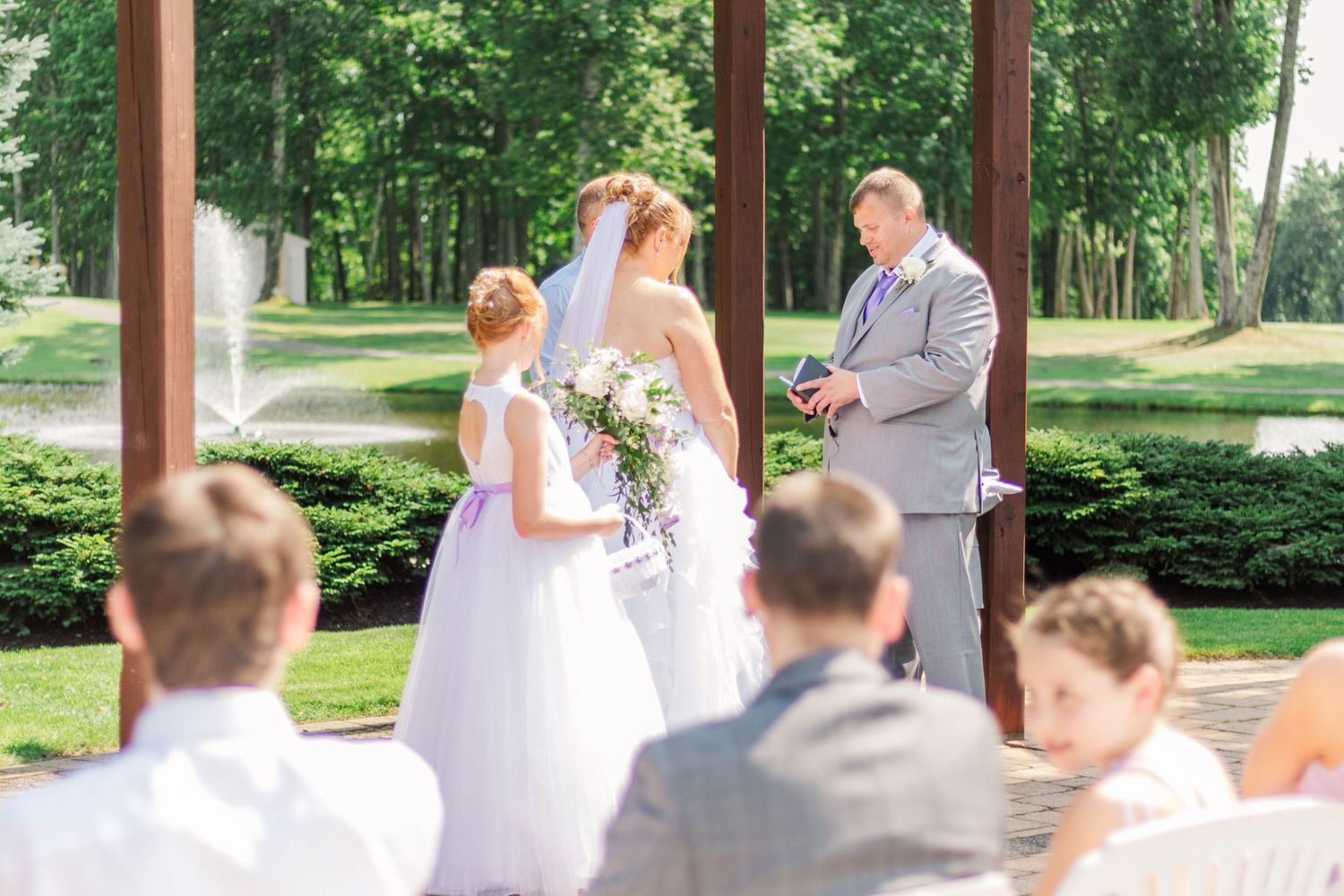 Outdoor wedding ceremony under wooden pergola with guests seated on white chairs.