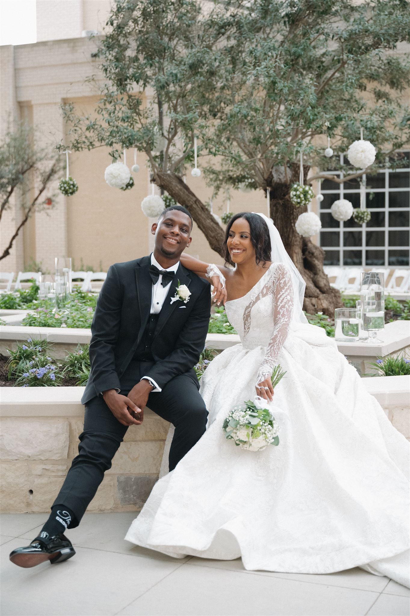 A happy couple shares a joyful moment at their outdoor wedding, with hanging lanterns and olive trees in the background.