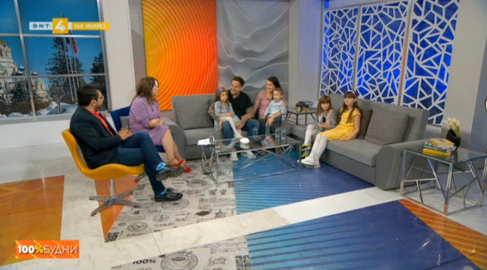 Talk show guests and hosts engage in conversation on a modern studio set with blue and orange decor.