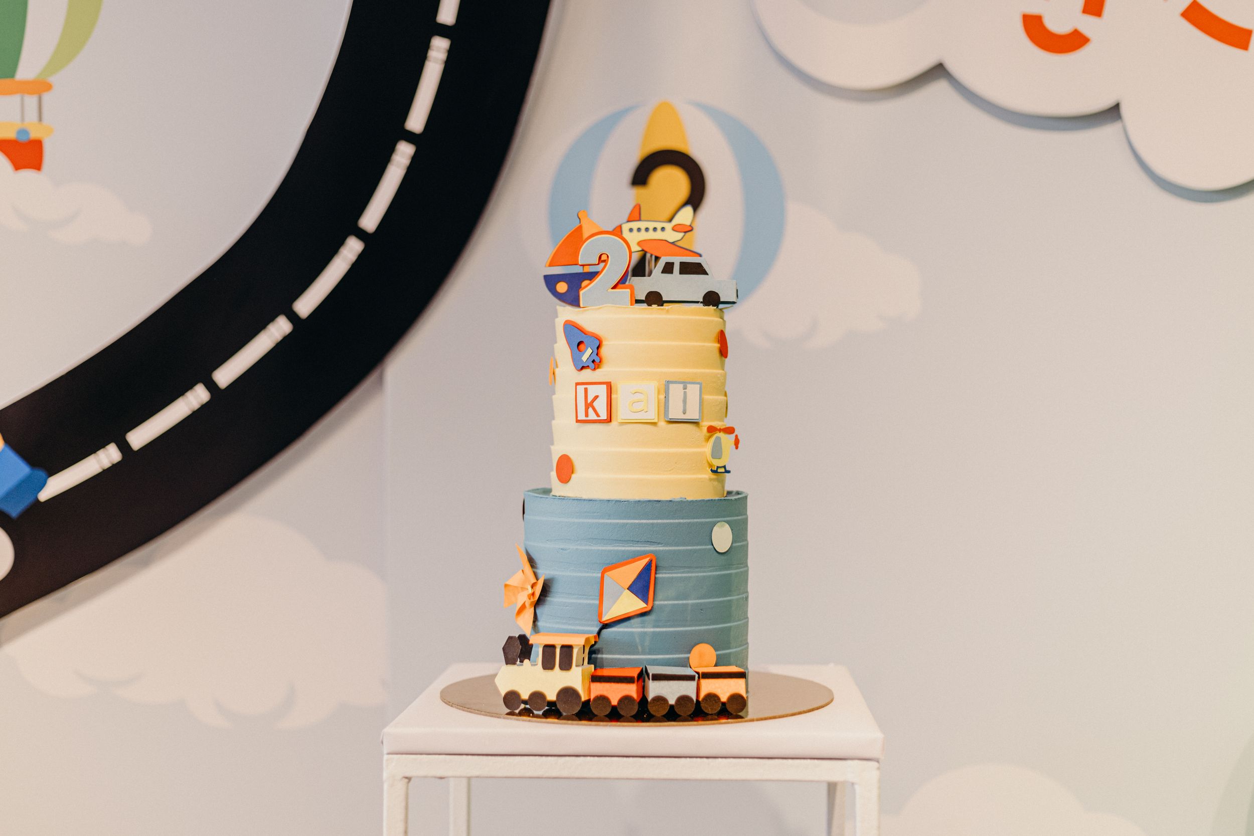 A whimsical fondant cake decorated with a train track, clouds, and toy train figures on multiple tiers.