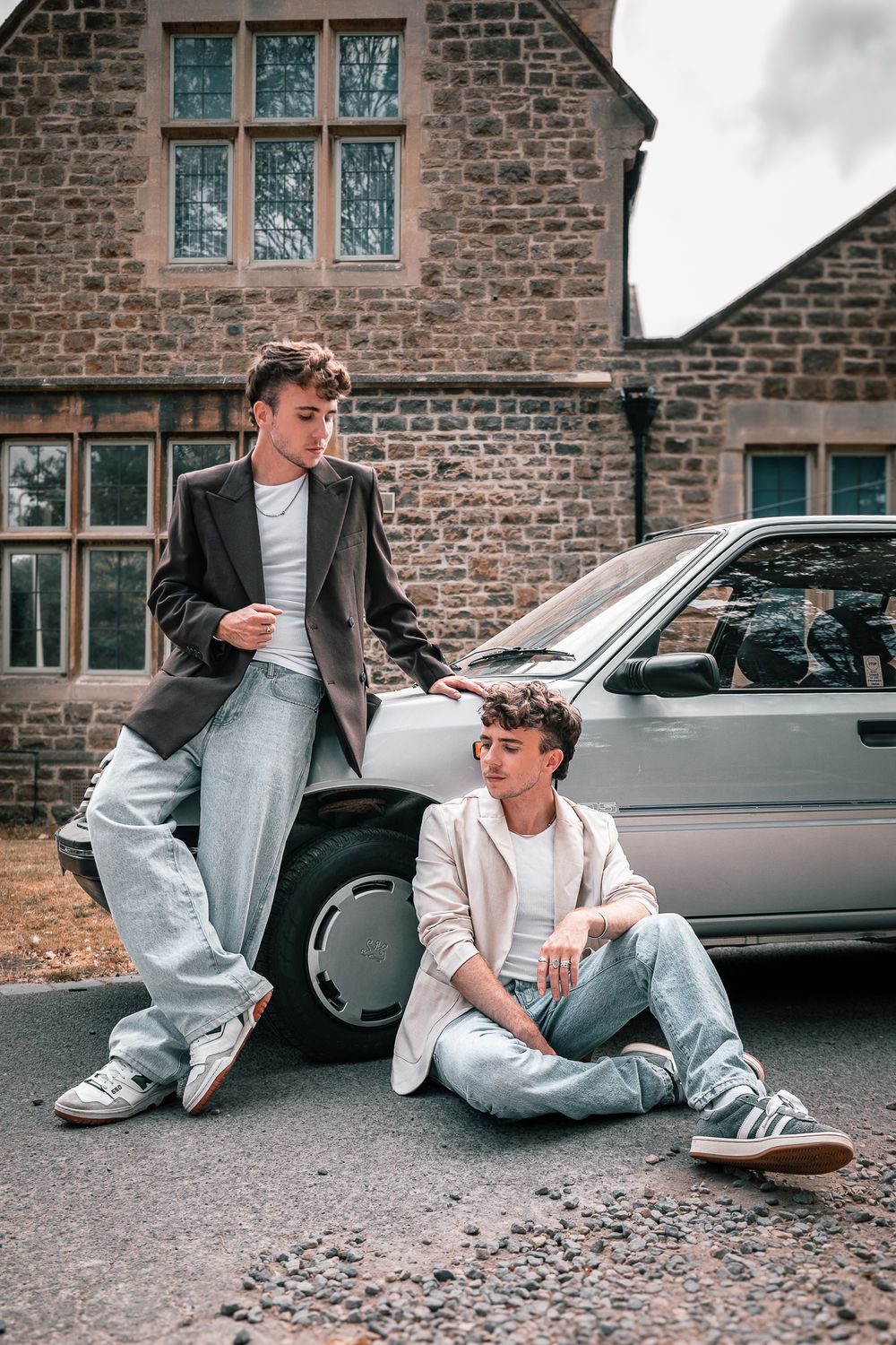 Two stylish young men posing near a silver car in front of a stone building, wearing casual jeans and blazers.