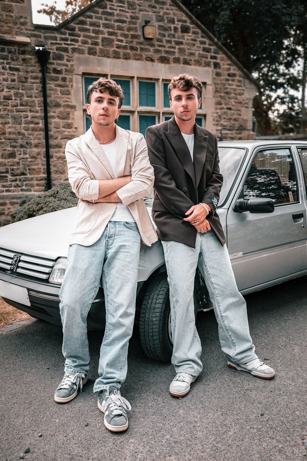Two young men in blazers and jeans leaning against a silver car outside a stone building.