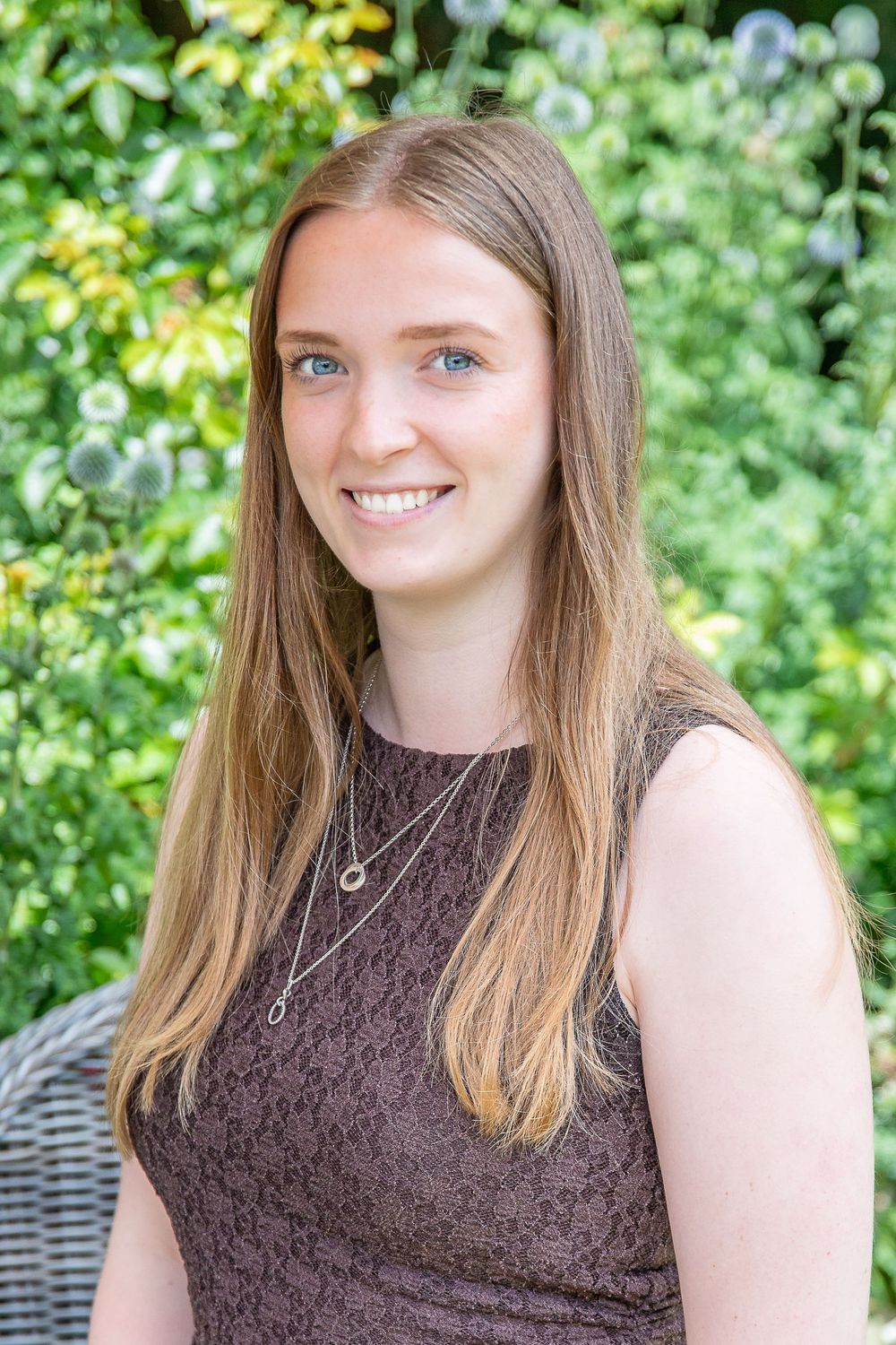 Young woman with long blonde hair and blue eyes smiling outdoors in a garden wearing a brown sleeveless top.