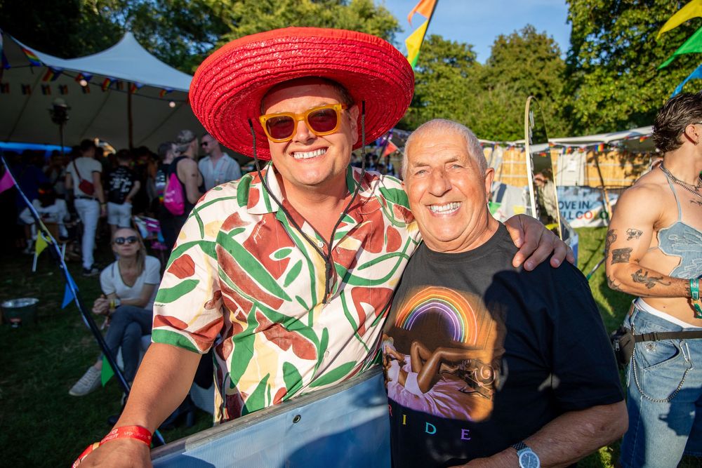 Two men smiling at a colorful outdoor festival, one wearing a red sombrero and tropical shirt.
