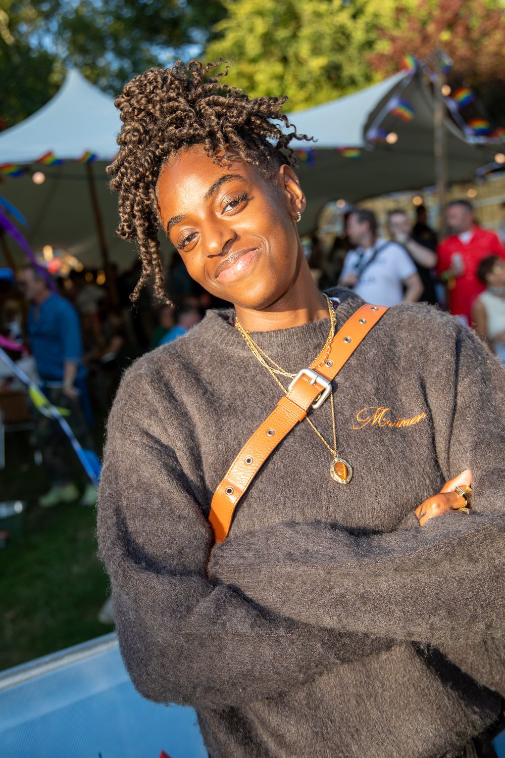 Young woman with locs, wearing gray sweater and gold necklace, arms crossed, smiling at outdoor festival.