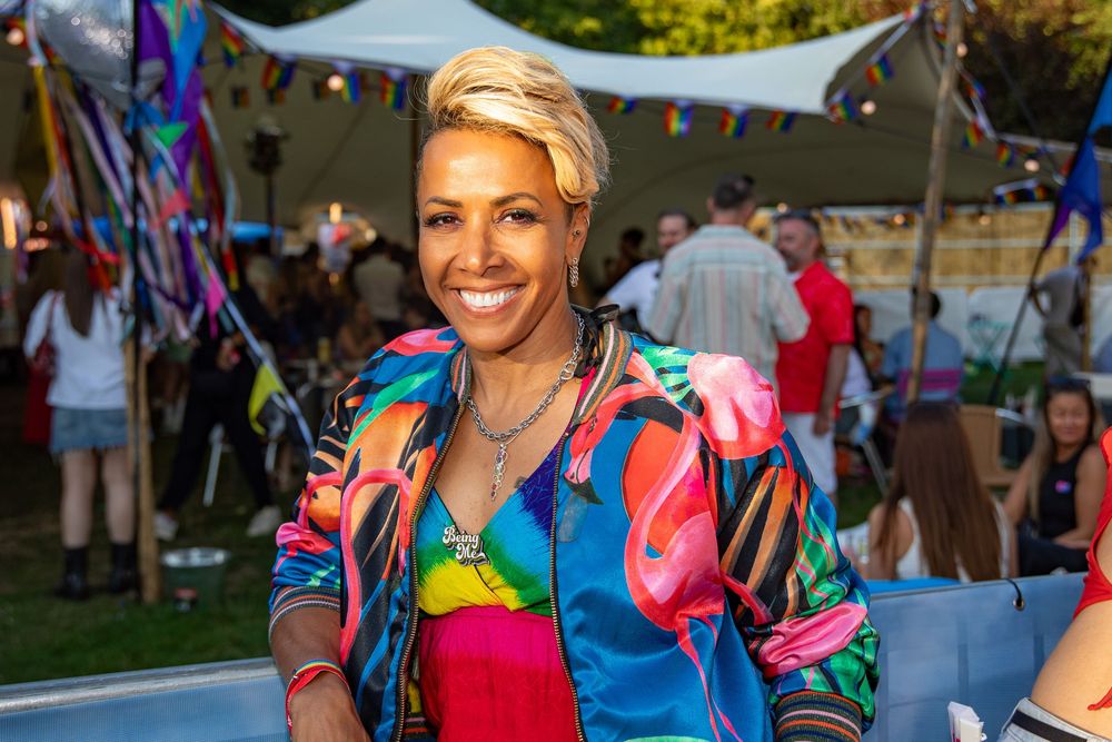 Smiling woman with short blonde hair wearing a colorful rainbow jacket at an outdoor festival event.