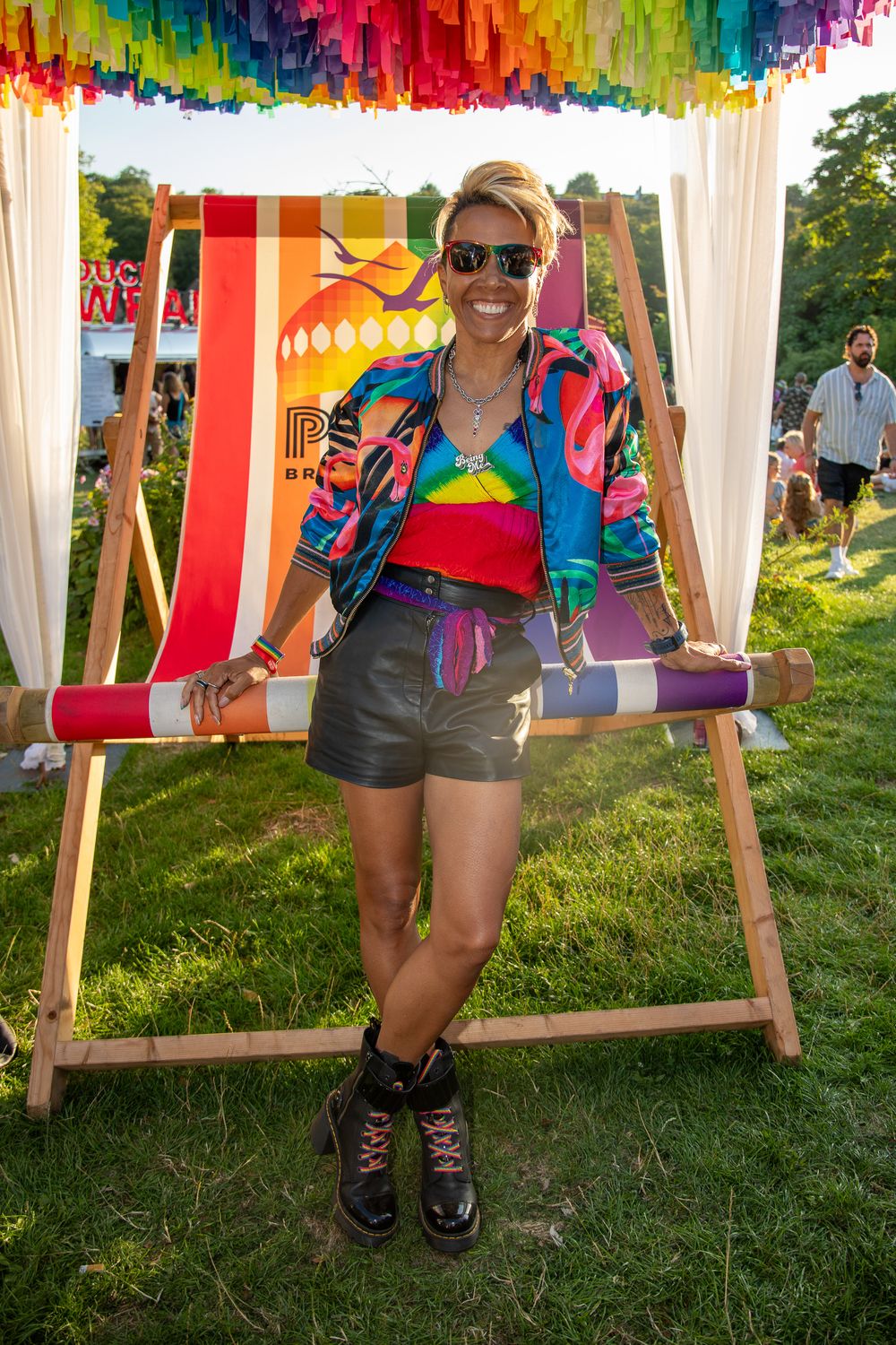 Woman in colorful rainbow jacket and shorts posing by giant deckchair at outdoor festival with rainbow decorations.
