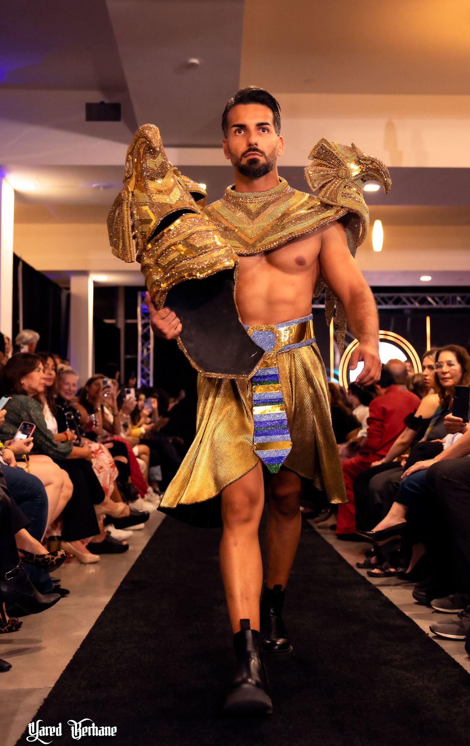 Muscular model wearing elaborate gold Egyptian-inspired costume walks runway at fashion show.