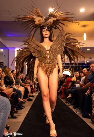 Model presents avant-garde brown feathered bodysuit with dramatic wing-like headpiece on runway.