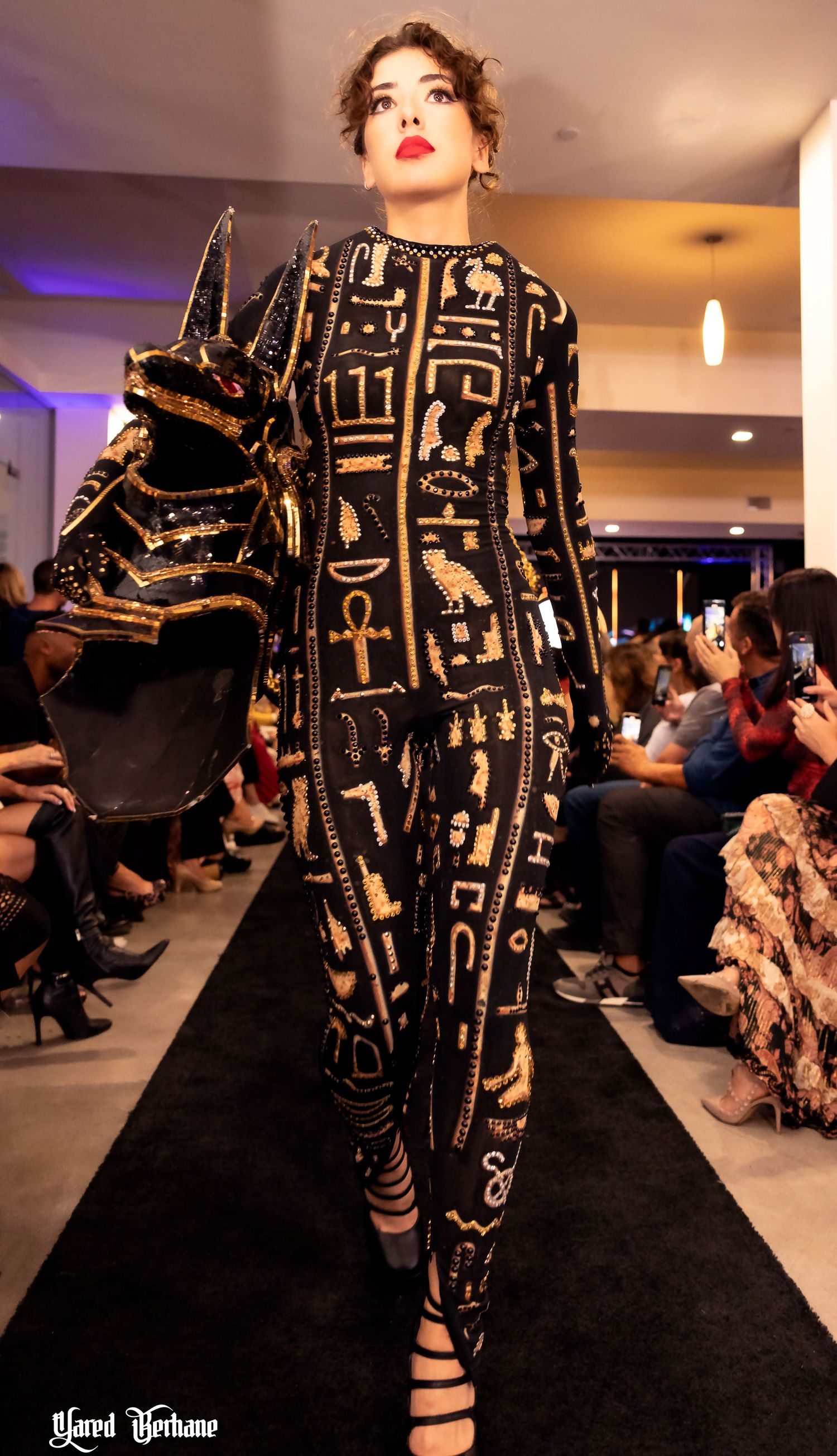 Model showcases Egyptian-inspired black and gold jumpsuit with hieroglyphic patterns on fashion runway.