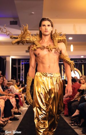 Model walks runway in gold harem pants with decorative golden branches across shoulders.