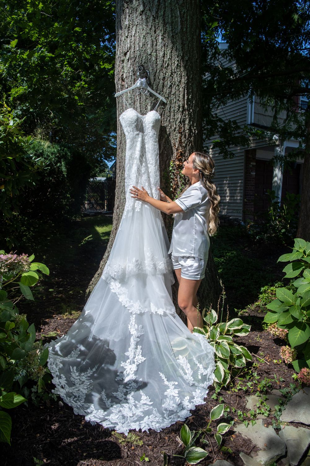 Bride in white pajamas admires her lace wedding gown hanging on a tree outdoors.