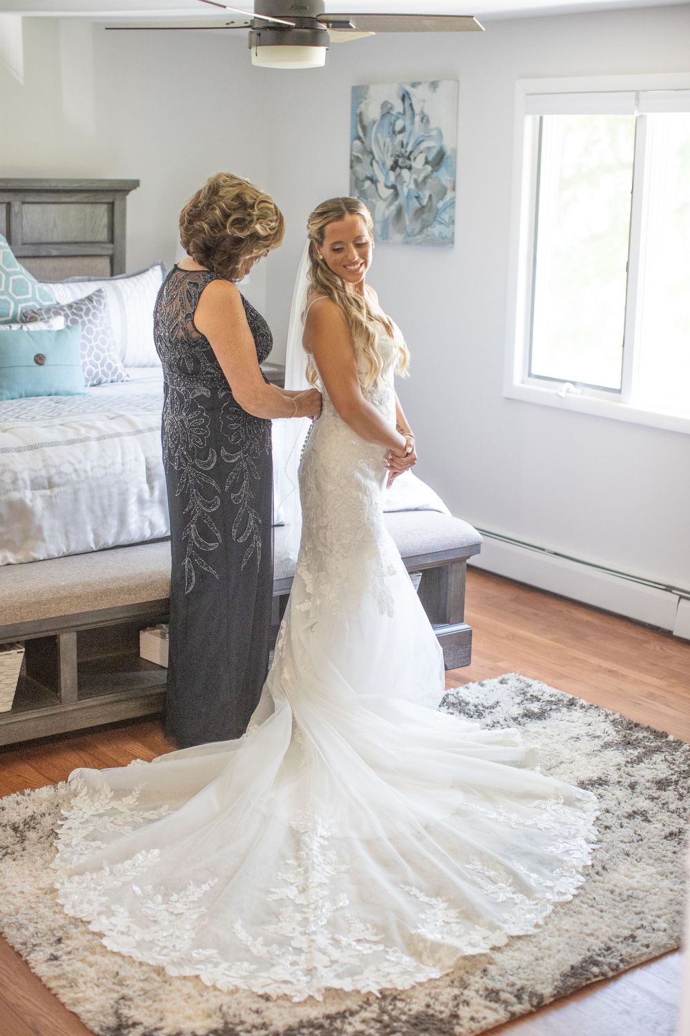 Mother helps bride button elegant lace wedding gown with long train in bedroom.