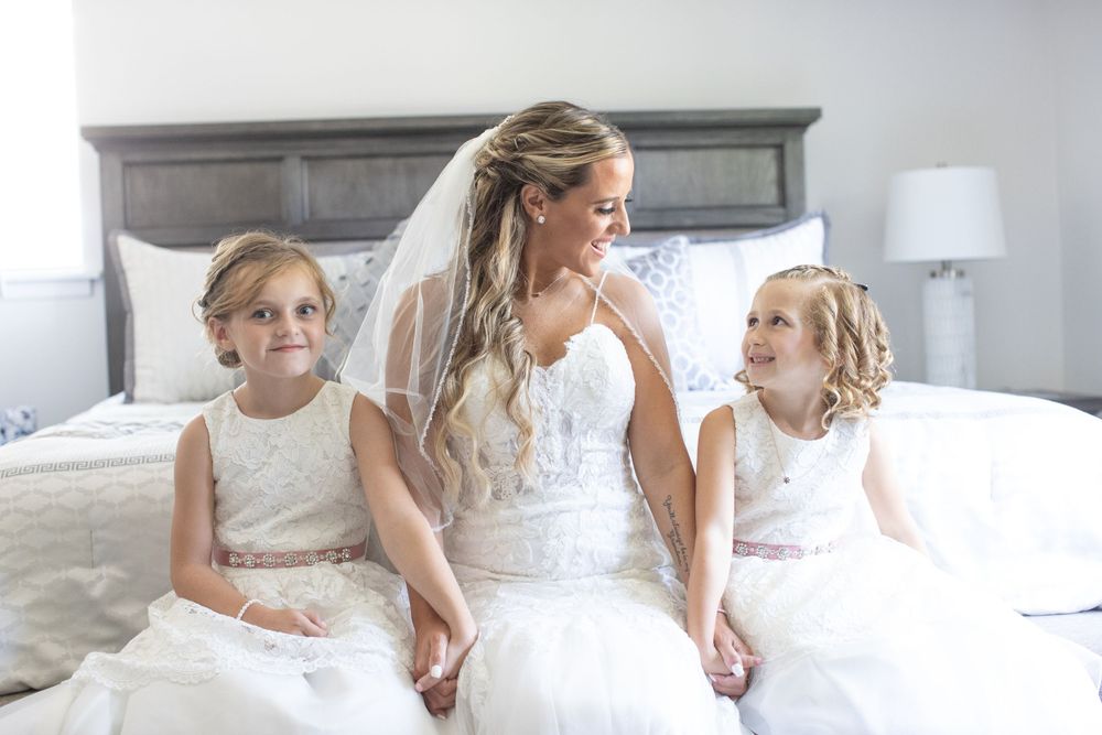 Bride in white lace wedding gown sits on bed with two young flower girls in white dresses.