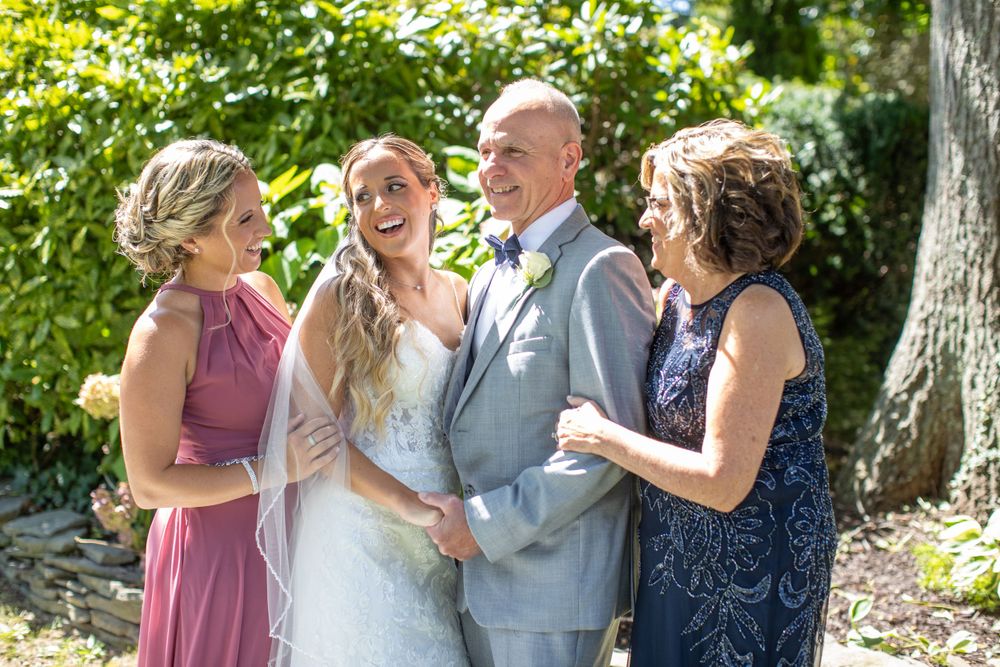 Bride in white gown laughs joyfully with family members outdoors on wedding day.
