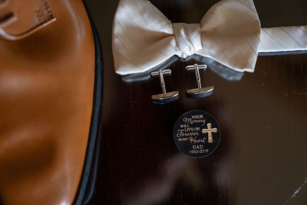 Wedding day groom details featuring cufflinks, white bow tie, leather shoes, and an engraved keepsake coin.