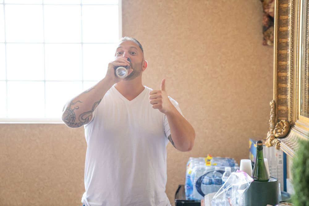 Groomsman in white t-shirt drinks a can and gives a thumbs up while getting ready for the wedding celebration.