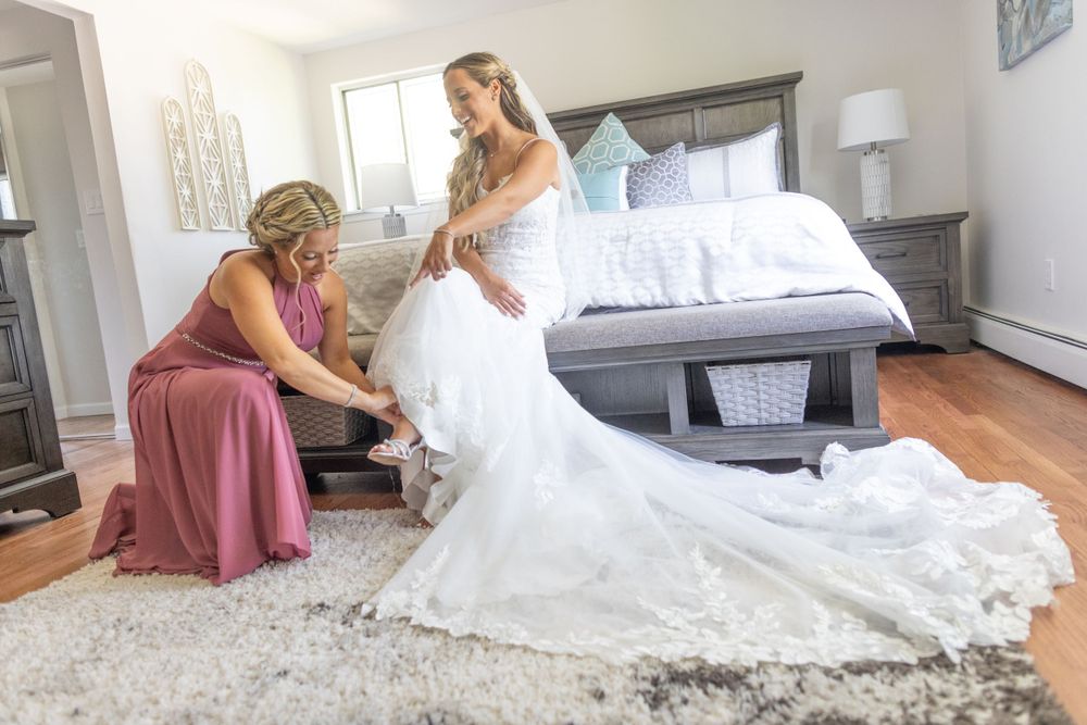 Bridesmaid in pink dress helping blonde bride with shoes in elegant bedroom before wedding.