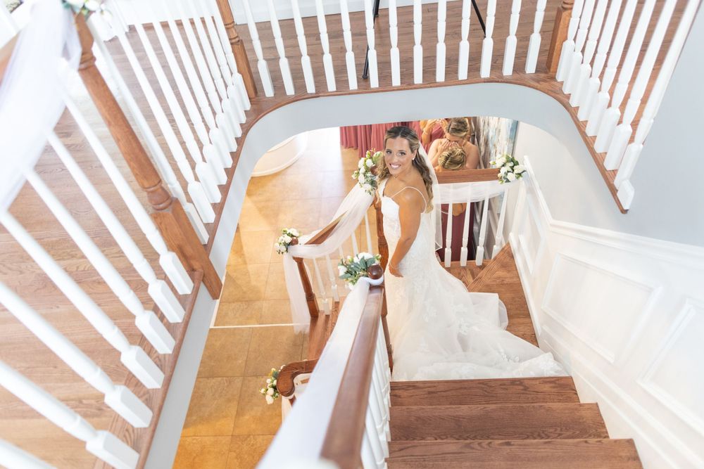 Elegant bride in white gown descending grand wooden staircase holding bridal bouquet overhead view.