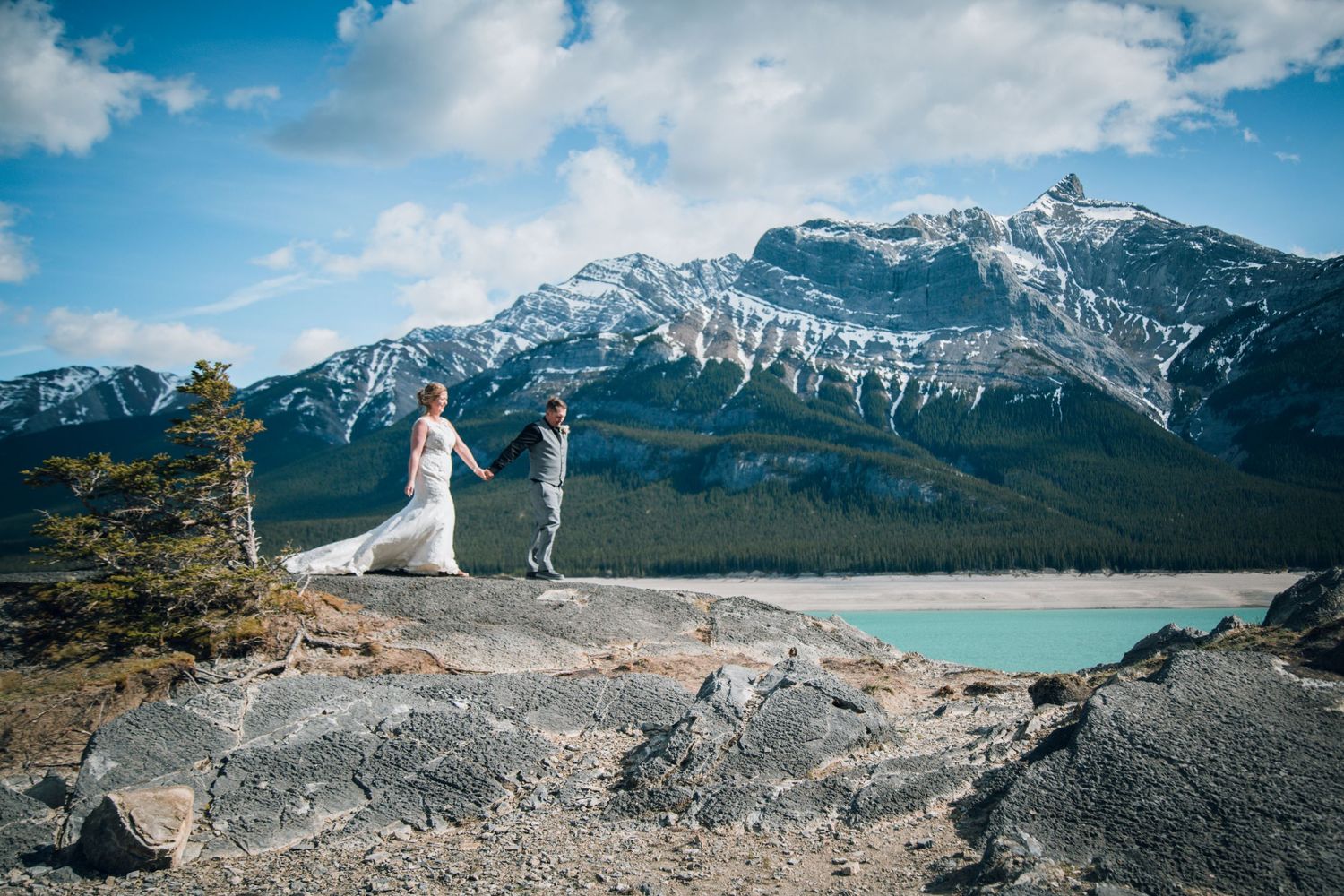 Wedding couple poses on rocky overlook with snow-capped mountains and turquoise lake in background.