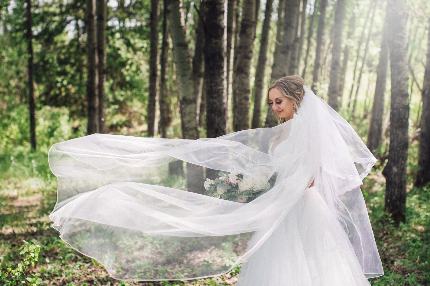 Flowing white wedding veil catches the breeze in a sunlit forest setting.
