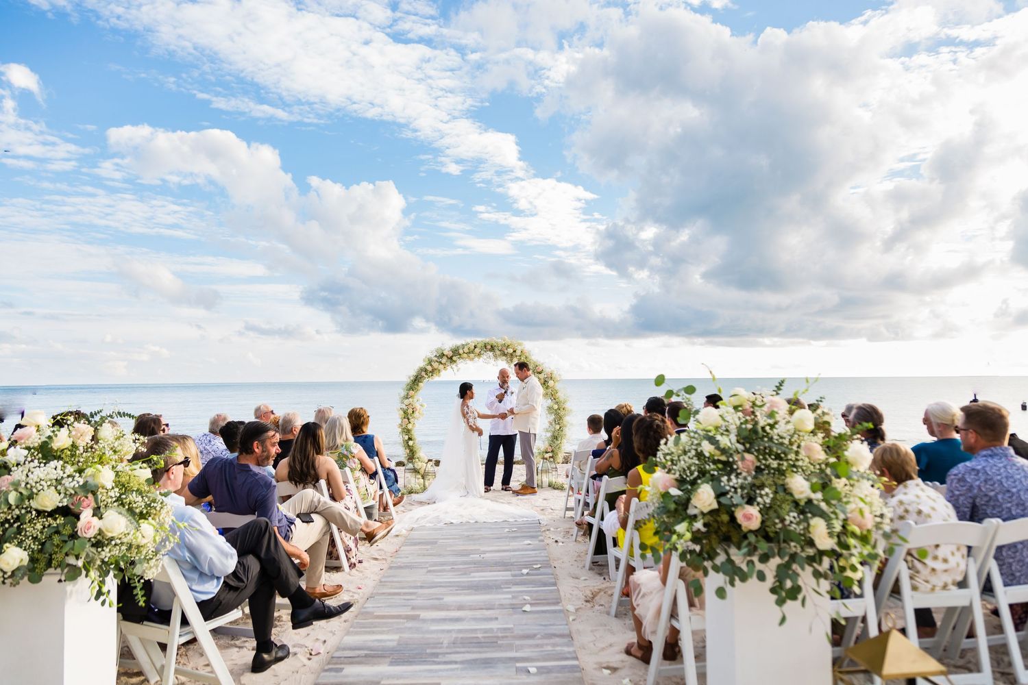 Outdoor beach wedding ceremony setup with white floral arch and guest seating.