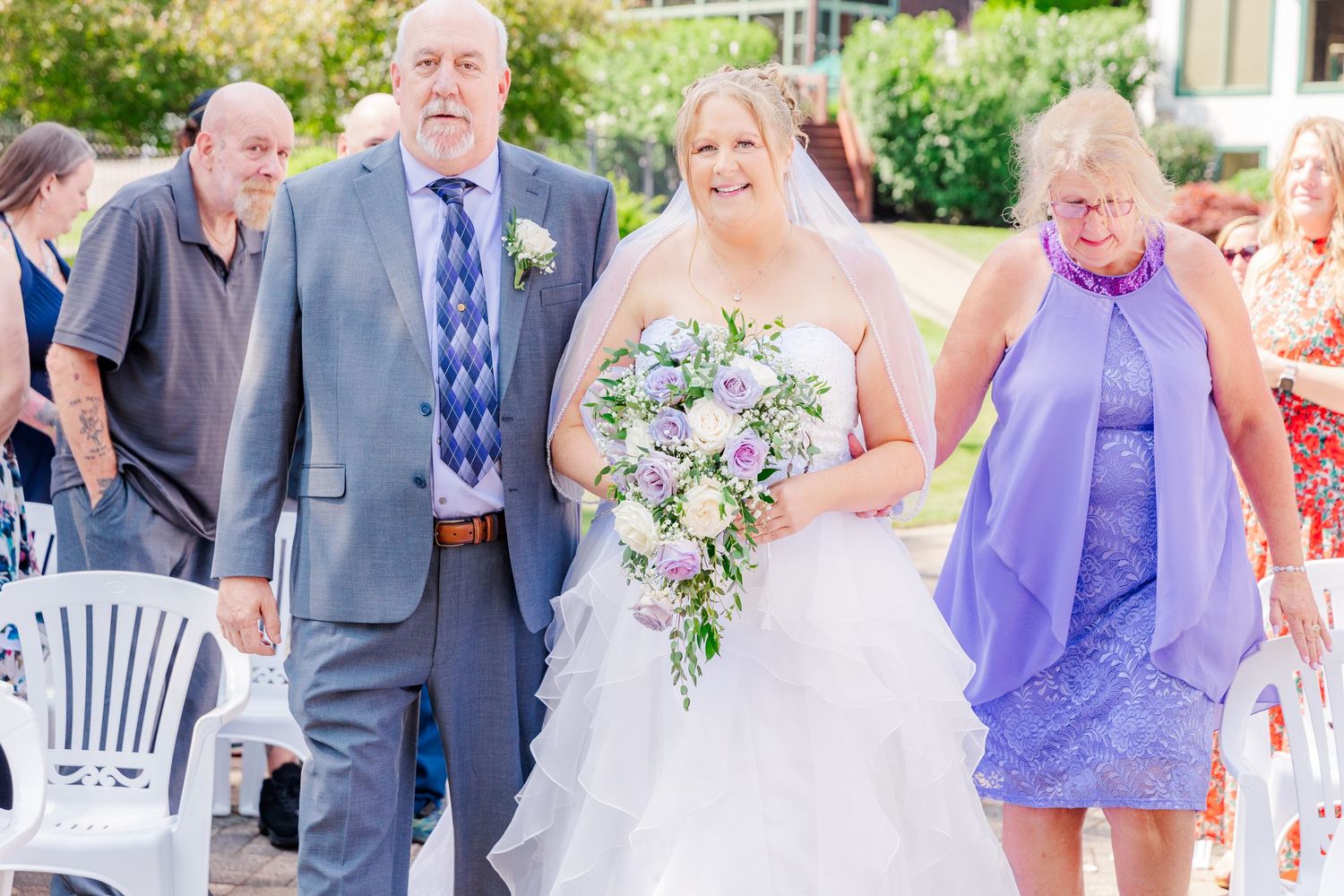 A wedding ceremony outdoors with attendees in formal attire, featuring purple and gray color scheme and floral bouquet.