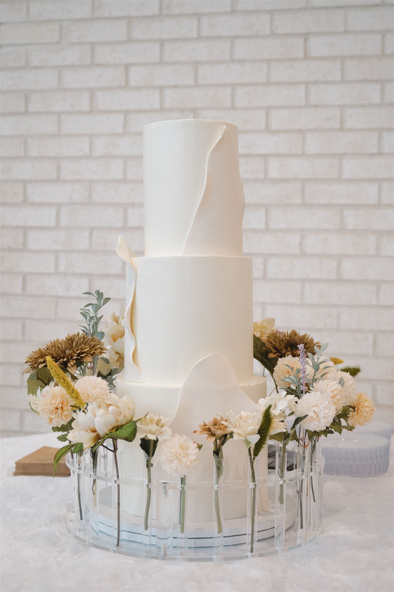 Elegant three-tier white wedding cake decorated with fresh flowers and greenery on a glass stand against a brick wall.
