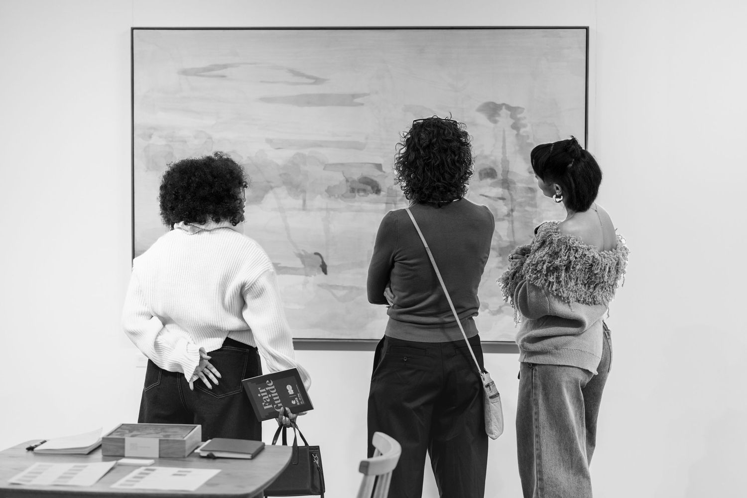 Three people stand observing artwork displayed on a gallery wall in a black and white photograph.