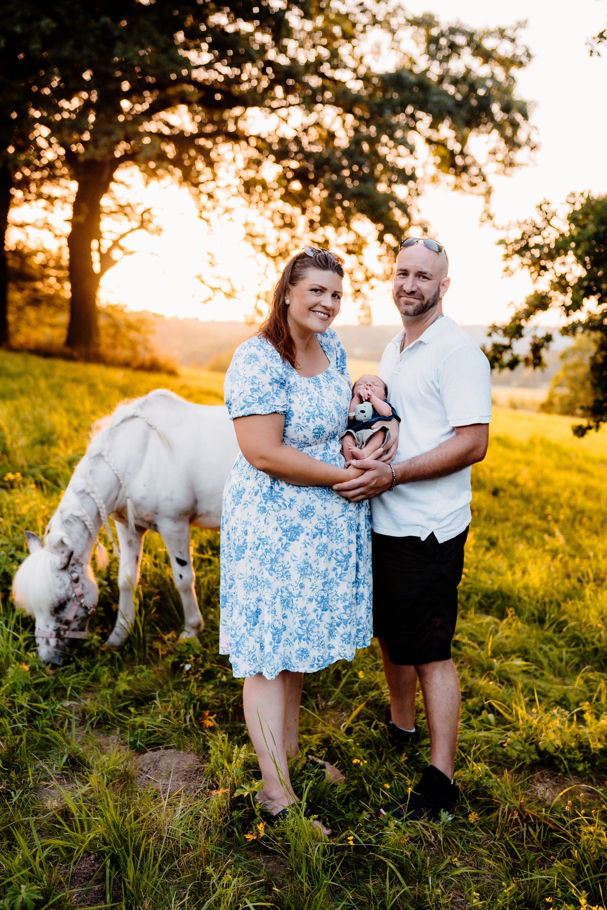 Couple in blue dress and white shirt share joyful moments with their fluffy white dog in a golden sunset meadow.