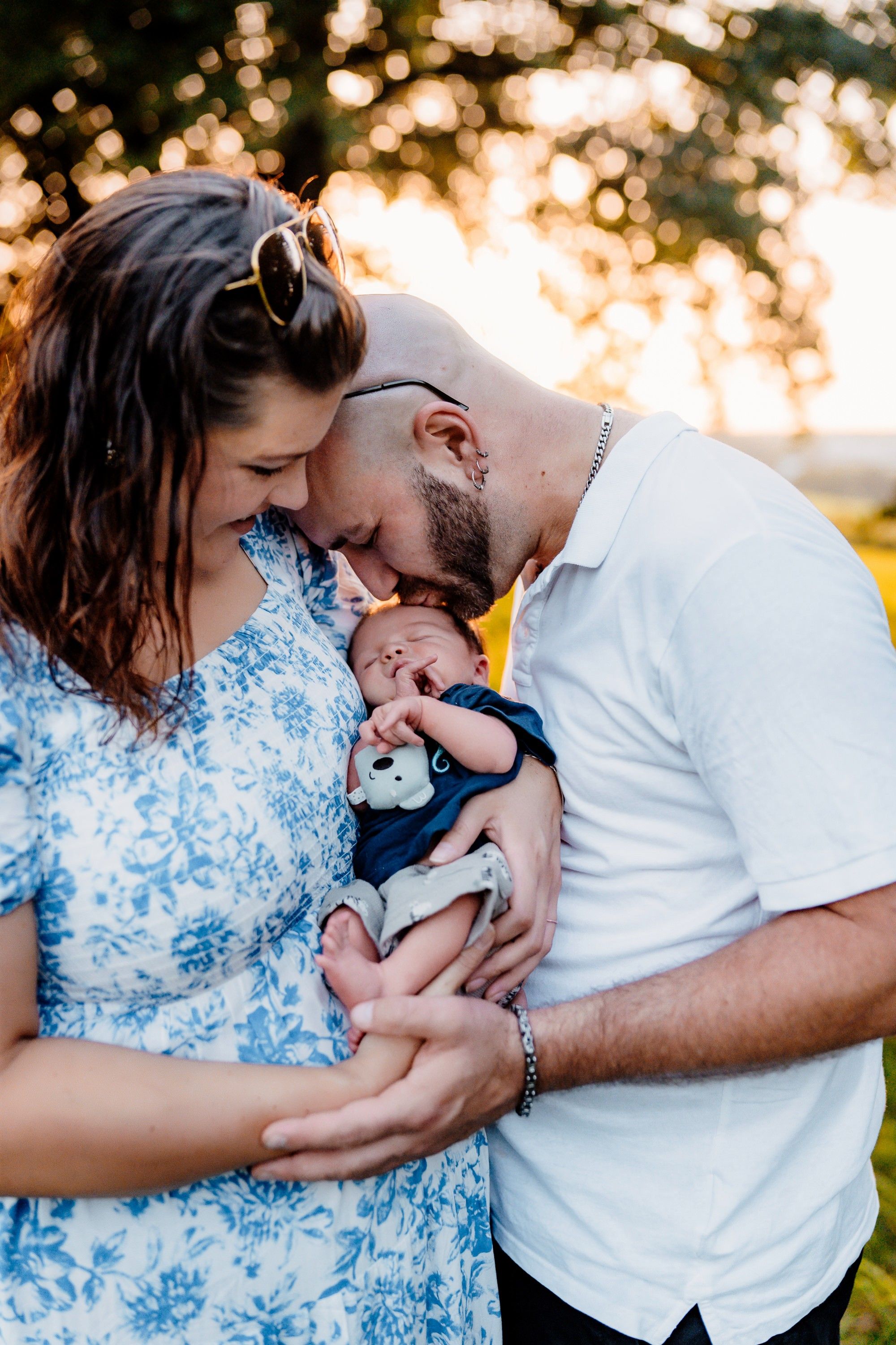 A couple holds their newborn baby outdoors during golden hour with sunlight bokeh in the background.