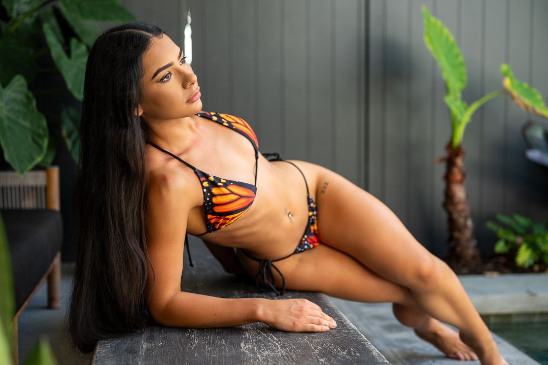 A woman in a butterfly print bikini poses on a stone surface near tropical plants in an outdoor setting.