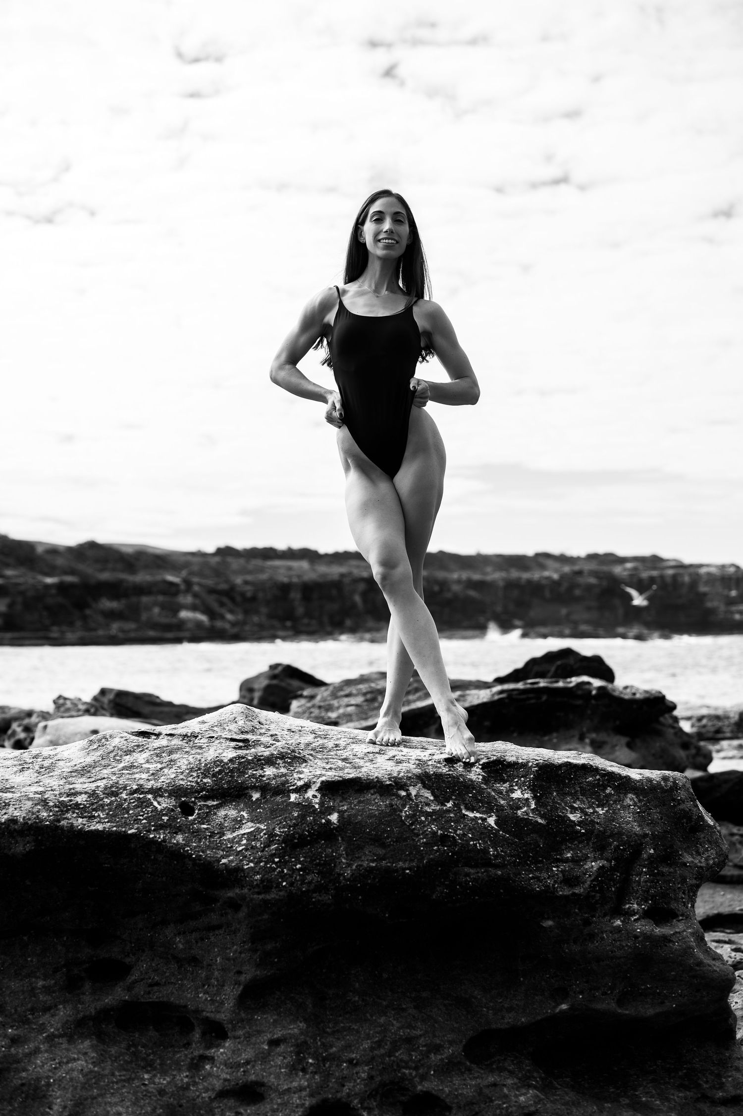 Black and white photo of a ballet dancer in a leotard striking an elegant pose on coastal rocks by the ocean.