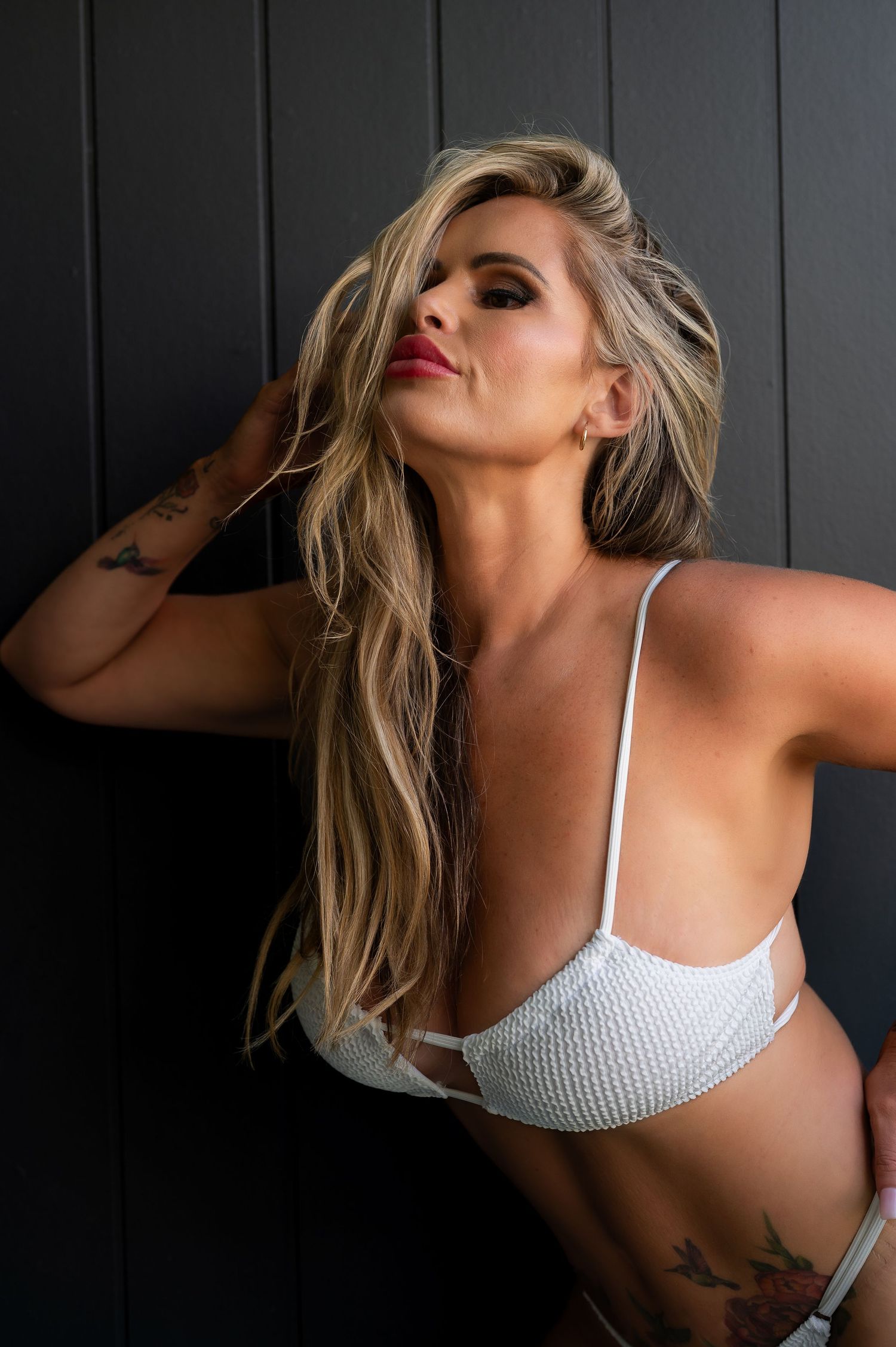 Fashion model in white knit bikini top poses against dark wall with flowing blonde hair and tattoos.