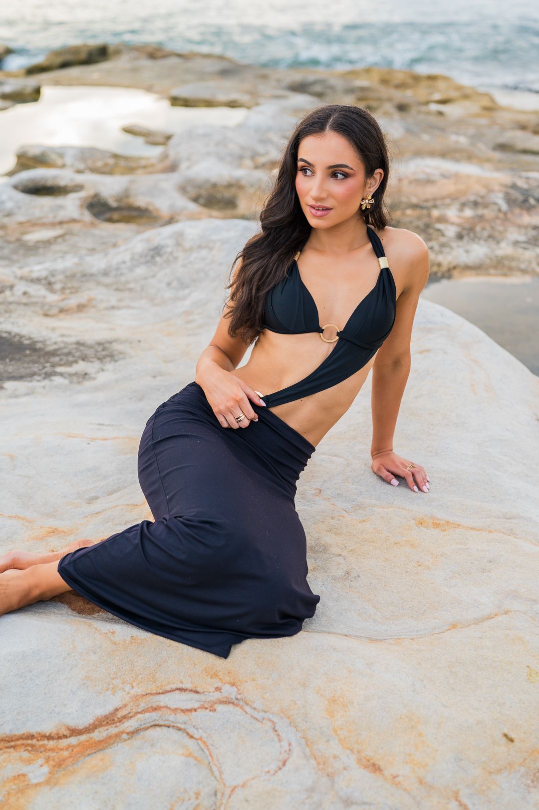 Model in black bikini top and flowing skirt poses elegantly on coastal rocks near ocean.