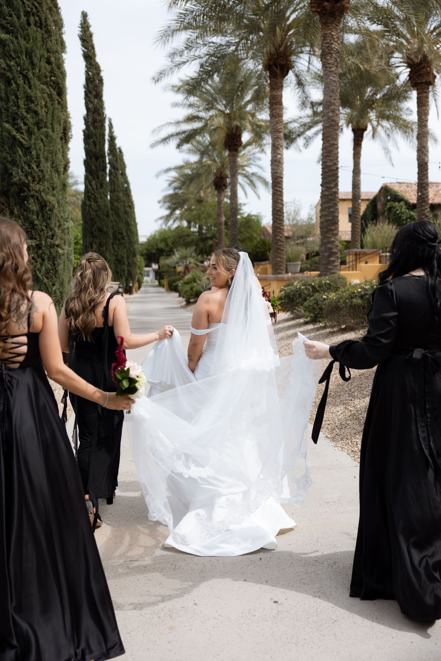 Bride in white gown and veil walks with bridesmaids in black dresses along palm tree-lined path.