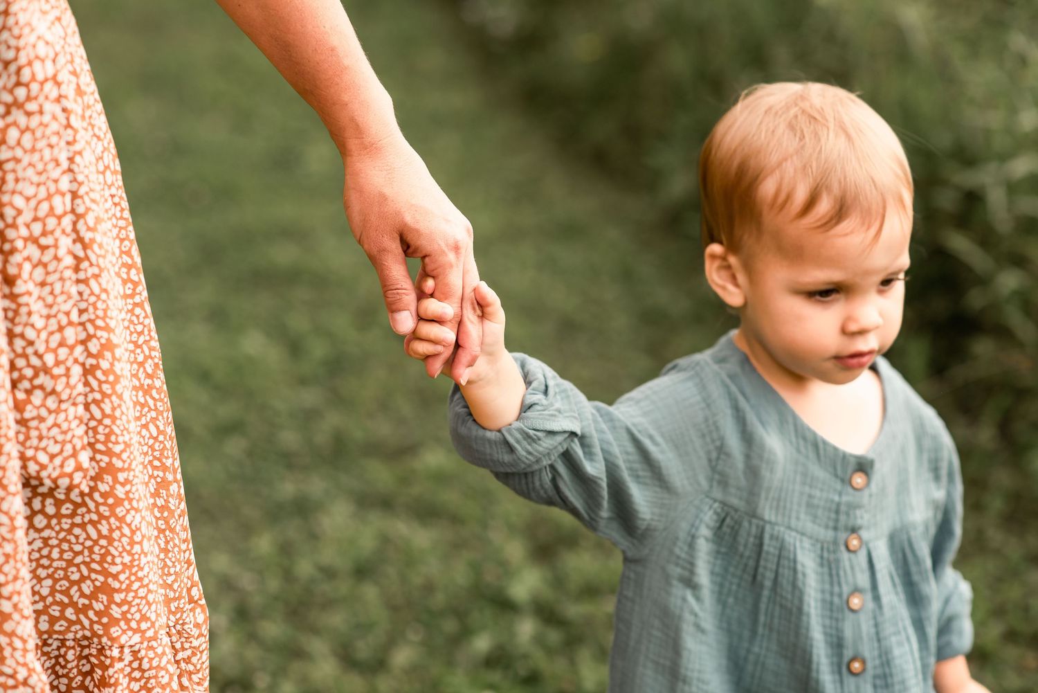Small child in grey outfit holding adult's hand while walking outdoors.