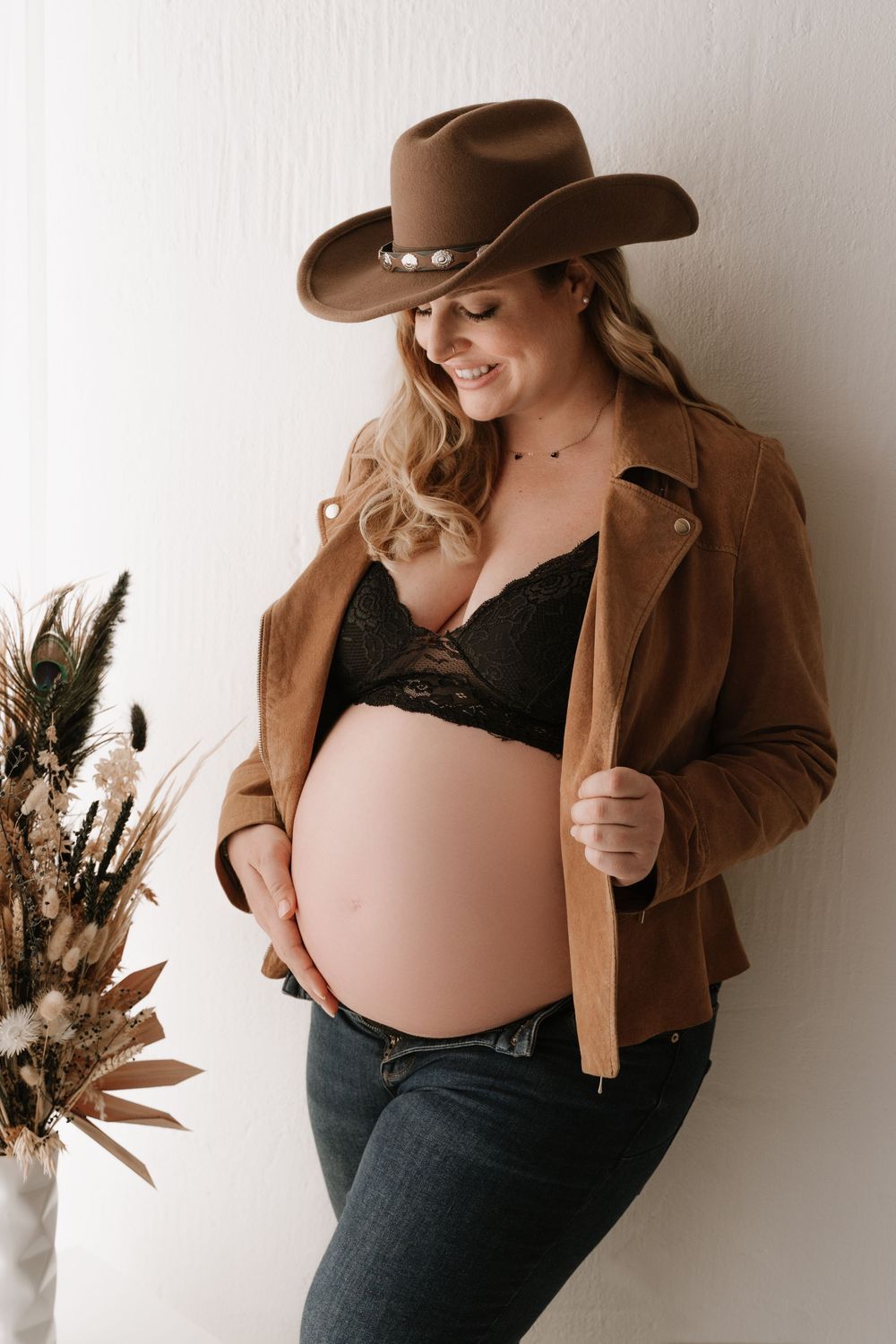 Smiling pregnant woman in cowboy hat and brown jacket proudly showing baby bump in studio.