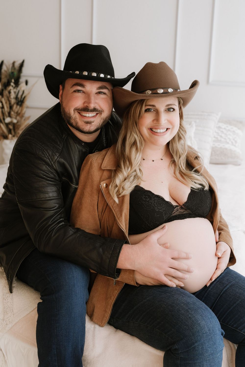 Smiling couple wearing cowboy hats sitting together, man embracing pregnant woman showing baby bump.