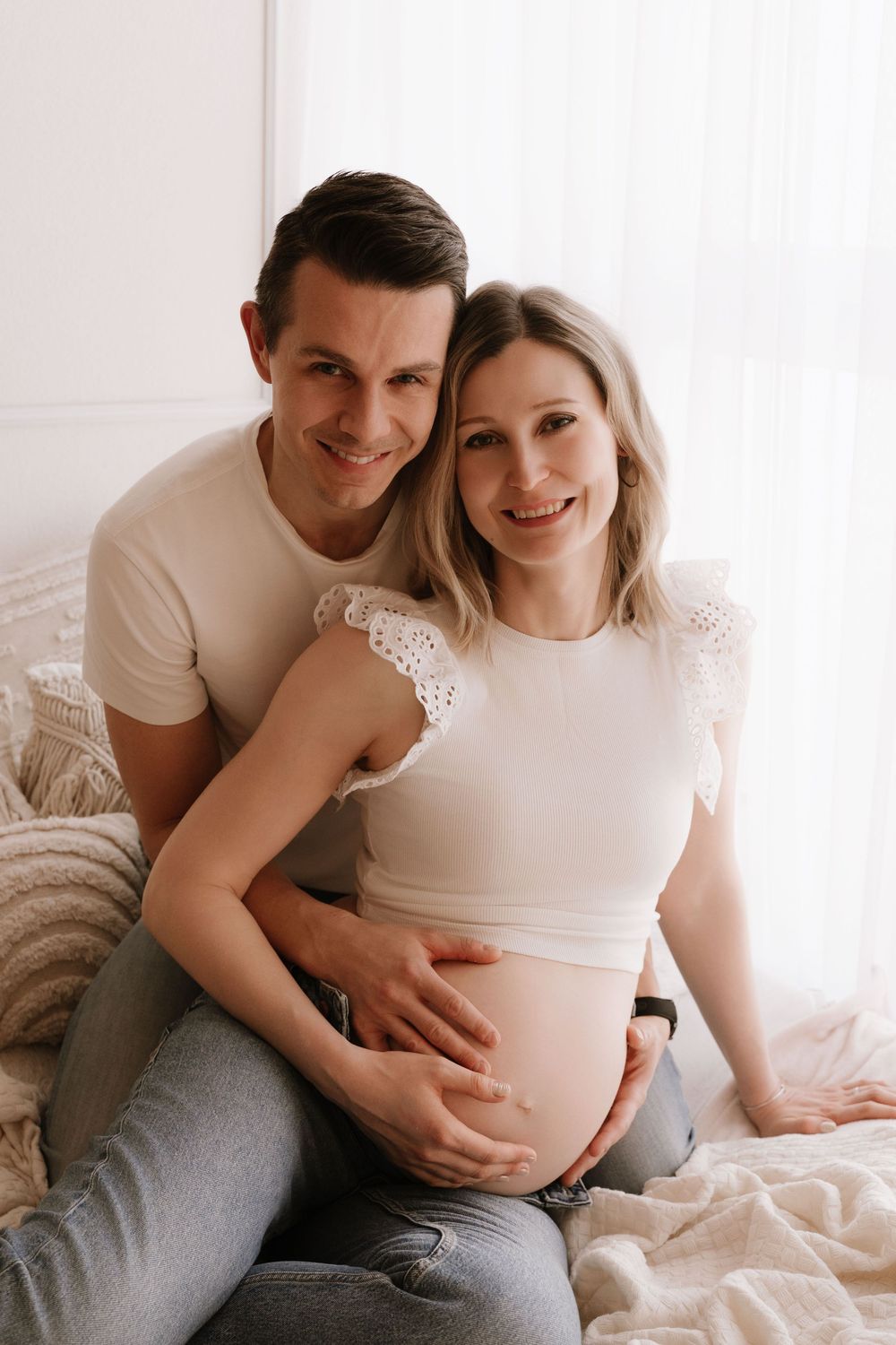Smiling couple expecting a baby, man embracing pregnant woman from behind on a cozy bed.