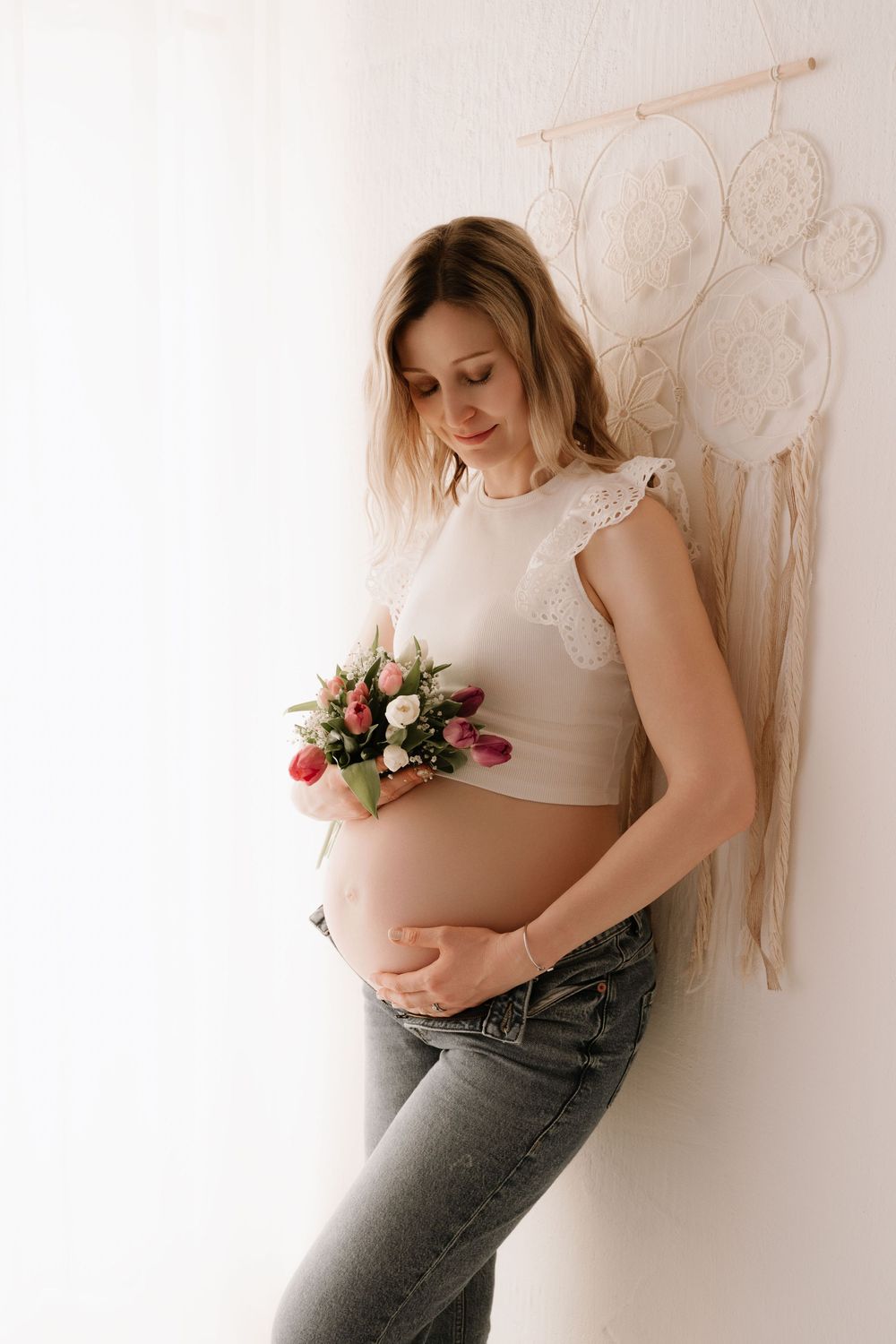 Pregnant woman in white crop top and jeans holding a bouquet of flowers against a white wall.