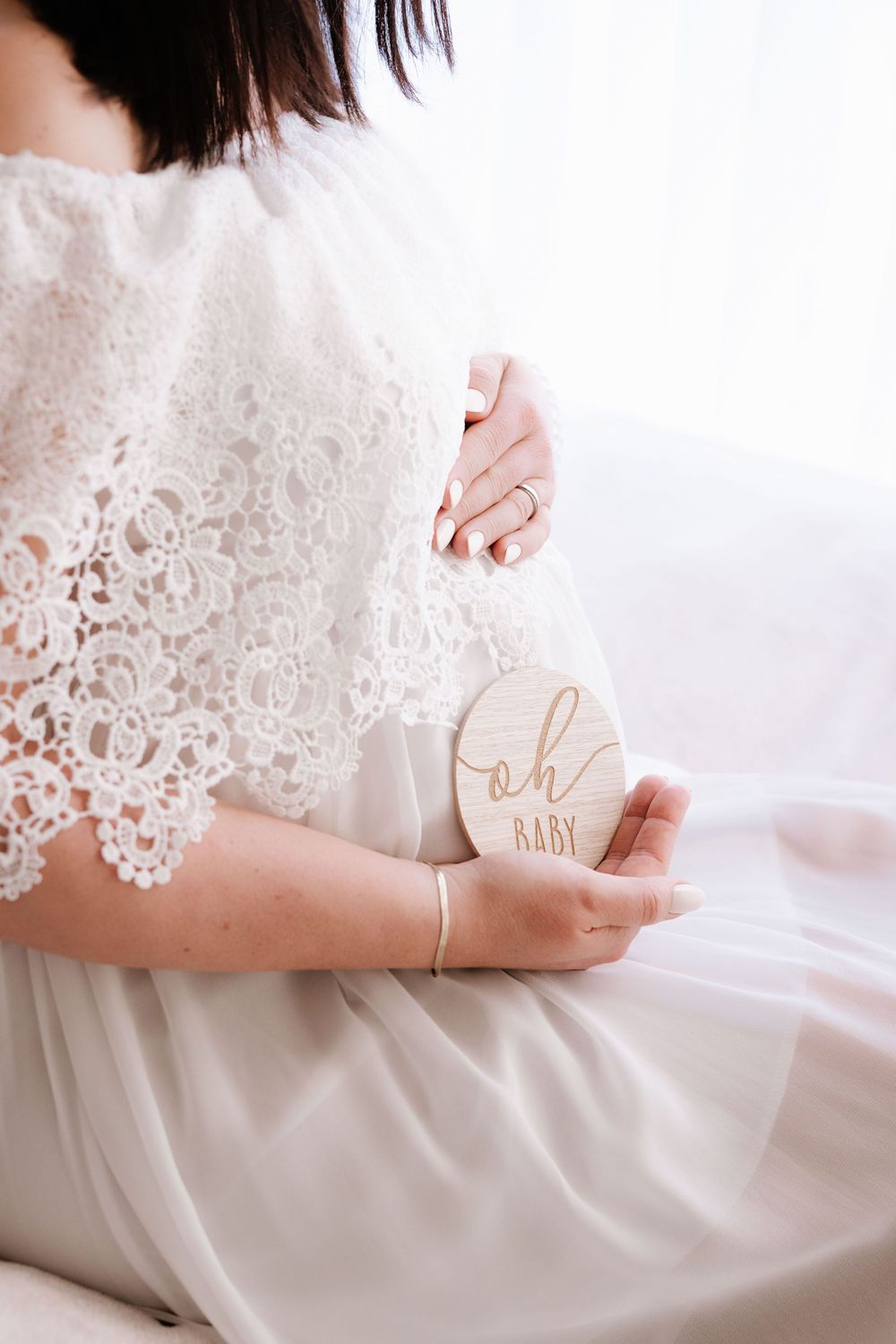 Pregnant woman in white lace dress holding a wooden oh baby disc while cradling her belly.
