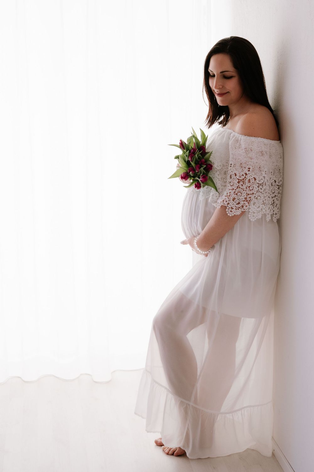 Pregnant woman in white lace off-shoulder gown holding red tulips while leaning against a wall.