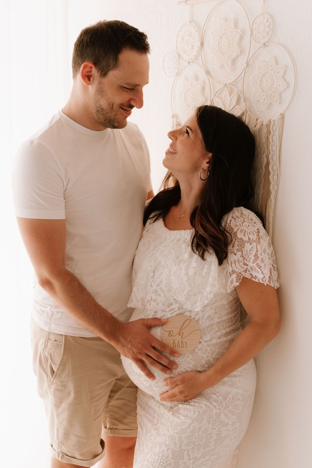 Happy couple smiling at each other, man holding pregnant woman's belly with wooden sign.