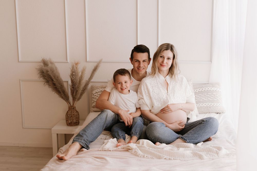 Happy family with pregnant mother, father, and young son sitting together on a bed in a bright white room.