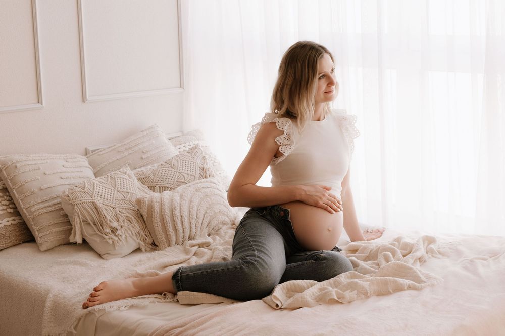 Pregnant woman in white top and grey jeans sitting on boho bed gazing out window.