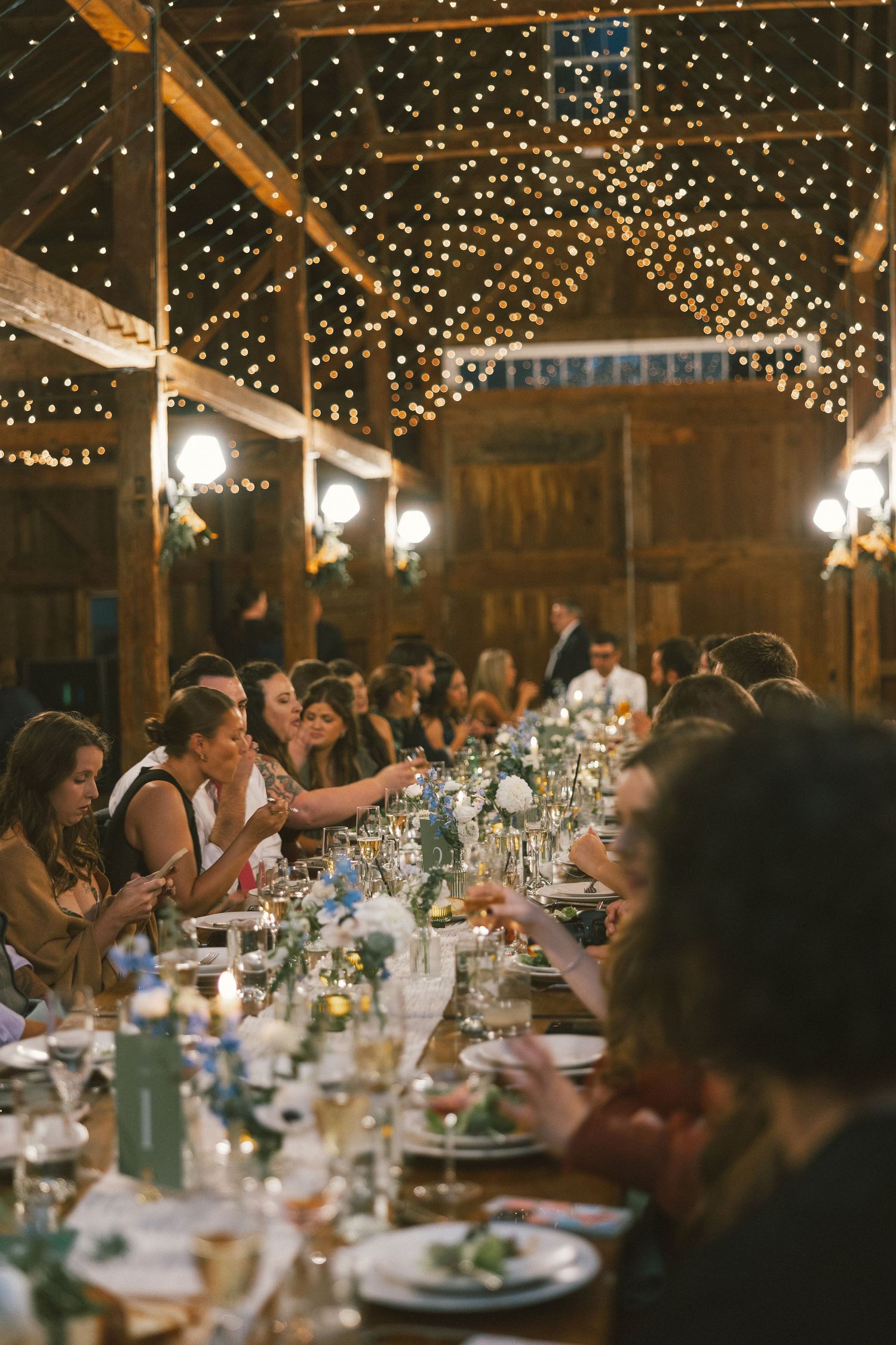 Wedding reception in a rustic barn with twinkling lights and long tables set for dinner.