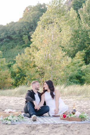 Couple sitting on a blanket on the beach surrounded by flowers and champagne for a picnic.